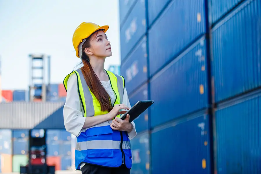 a young female shipping yard employee looking up at some containers while holding a clipboard