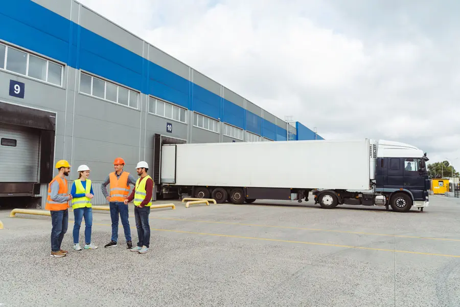 a group of workers standing by semi-truck while it is being loaded with shipping pallets